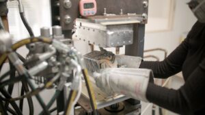 Metal sheet being shaped in a factory stamping press by a worker wearing safety gloves.
